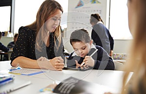 Young female teacher working with a Down syndrome schoolboy sitting at desk using a tablet computer and stylus in a primary school