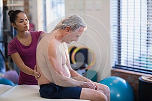 Young female therapist giving back massage to senior male patient sitting on bed