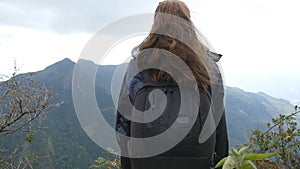 Young female tourist with backpack enjoying beautiful view in the mountains. Woman traveler in raincoat standing on the