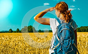 Young female tourist with backpack looking into the distance near a wheat field under the hot summer sky.