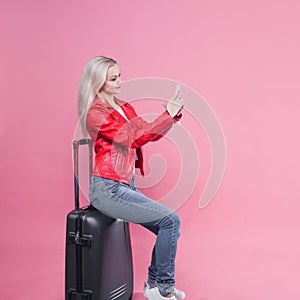 Young female tourist in a jacket and with Luggage on a pink background.