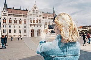 Young female tourist takes pictures of Hungarian Parliament with her smartphone on beautiful sunny day. Shot from behind. European