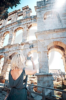 Young female tourist takes pictures of Pula Arena, Croatia with her smartphone on beautiful sunny day. Shot from behind. European
