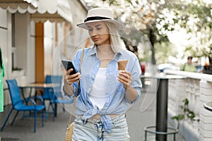 Young female tourist walking through the old town eating ice cream and using a smartphone to search for a map online