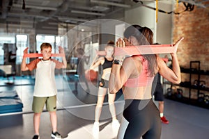 Young female trainer working out using resistance band in gym together with kids, teenagers. Sport, healthy lifestyle