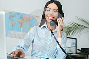 Young female travel agent consultant in tour agency answer phone call browsing laptop