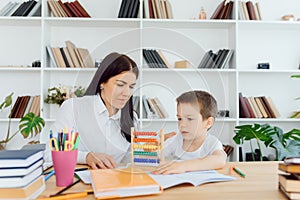 Young female tutor helping little elementary school boy with homework during individual lesson at home