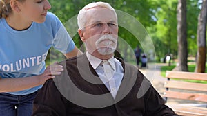 Young female volunteer walking in hospital garden with old man in wheelchair