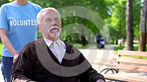 Young female volunteer walking in hospital garden with old man in wheelchair