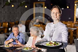 Young female waitress stands with order tray in spacious guest room in restaurant.