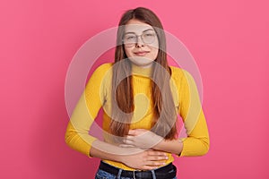 Young female wearing casual yellow shirt and eyewear touching her stomach, suffering from abdominal pain, has period, posing