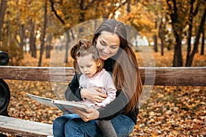Young female woman babysitter and toddler baby girl read book in autumn park. Happy family mom and toddler outdoors in