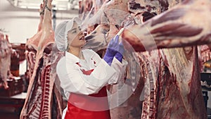 Young female worker of meat processing factory inspecting fresh raw meat hanging in cold storage room, monitoring