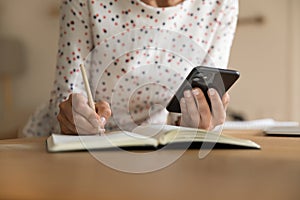 Young female writing information from smartphone screen to paper notebook