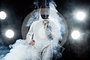 Young fencer athlete wearing fencing costume holding the sword and mask. Isolated on black background with lights photo