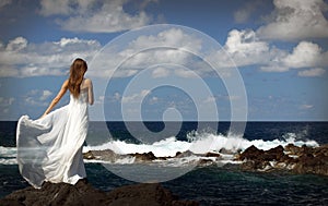 Young fiancee in light white wedding dress standing on windy sea roch shore on Sao Miguel island, Azores