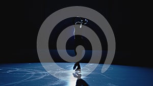 Young figure skating artist rotates on ice arena in dark against backdrop of bright blue spotlight. Silhouette of female