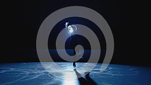 Young figure skating artist rotates on ice arena in dark against backdrop of bright blue spotlight. Silhouette of female