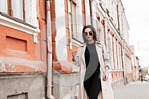 Young fine beautiful woman model in trendy sunglasses in fashionable beige trench coat walks near vintage building in city. Modern