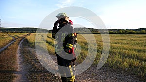 Young fireman in full equipment talking on walkie-talkie at countryside. Male firefighter speaking on radio