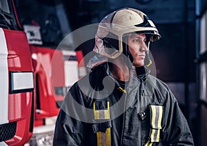 Young fireman wearing protective uniform standing next to a fire engine in a garage of a fire department