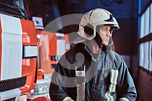 Young fireman wearing protective uniform standing next to a fire engine in a garage of a fire department