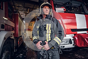 Young fireman wearing protective uniform standing next to a fire engine in a garage of a fire department