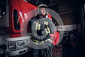 Young fireman wearing protective uniform standing next to a fire engine in a garage of a fire department