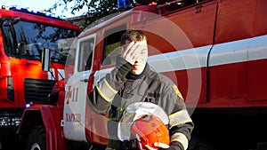 Young fireman wiping away the sweat on his face after hard work near fire engine. Portrait of tired male firefighter in