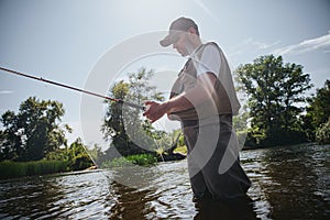 Young fisherman fishing on lake or river. Adult guy in fisherman`s clothes and cap holding rod in hands. Using lure for