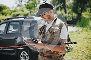 Young fisherman fishing on lake or river. Busy guy using smartphone. Stand at car and holding fishing rod before start