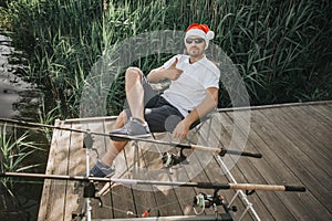 Young fisherman fishing on lake or river. Guy in festive christmas hat sitting alone on pier and posing on camera