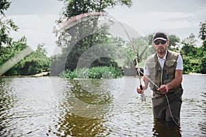 Young fisherman fishing on lake or river. Serious concentrated guy in fisherman`s clothes stand in river or lake water