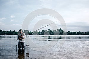 Young fisherman is standing barefeet in water and holding fly rod. He looks at it. Guy is working with spoon and bait