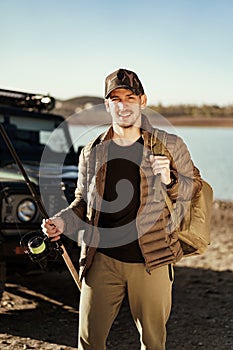 Young fisherman standing near his car and holding fishing equipment