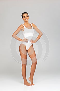 Young, fit and beautiful brunette woman in white swimsuit posing over grey background. Healthcare, diet, sport and
