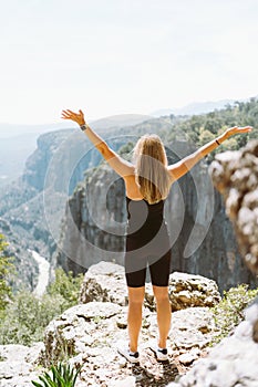 Young fit beautiful girl woman in black sporty slim outfit standing on edge of mountain cliff peak with hands up in air