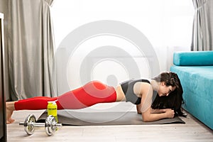 Young fit brunette woman wearing sportwear standing in plank after doing everyday warm up exercises with dumbbell on the floor at
