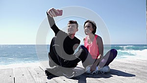 Young fit couple sitting near the beach holding phone and taking selfies posing. Happy boyfriend and girlfriend taking pictures. S