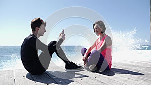 Young fit man sitting on the beach taking picture of his girlfriend with the phone. Strong waves splashing against the rocks