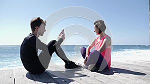 Young fit man sitting on the beach taking pictures of his girlfriend with the phone. Strong waves splashing against the rocks on t