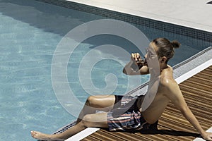 Young fit man sitting on the edge of the swimming pool relaxing while drinking a coke beverage and enjoying the weather