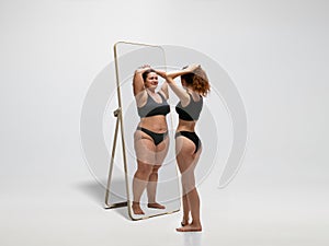 Young fit, slim woman looking at fat girl in mirror`s reflection on white background