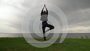 Young fit woman in black hoody practice yoga on coast near the lake or sea. Woman doing Tree pose Vrksasana Vriksasana