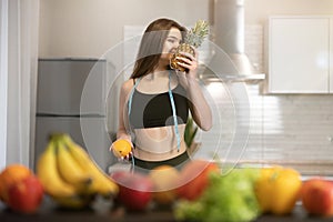 Young fit woman with centimeter round neck wearing black top and leggings smells pineapple standing in kitchen full of fruits