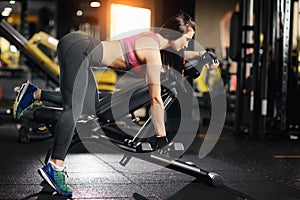 Young fit woman doing back dumbbell row exercises on a bench in the gym.