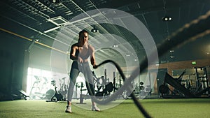Young fit woman doing different exercises on battle rope in well-equipped gym