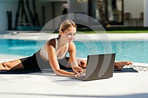 Young fit woman doing yoga stretching exercise outdoor near swimming pool in front of laptop.