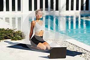 Young fit woman doing yoga stretching exercise outdoor near swimming pool in front of laptop.