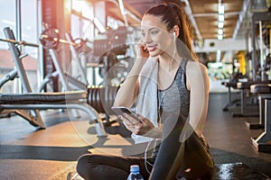 Young fit woman with earphones listening to music on her smart phone and taking a rest on fitness mat after gym workout.